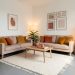 Salon d’appartement en location aux murs blancs et au sol gris, réchauffé par des coussins ocre, un tapis crème, des meubles en bois clair, des paniers tressés, des plantes et des lampes chaleureuses.