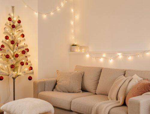 Petit salon lumineux décoré pour Noël avec un sapin mural pliable en pin clair, un canapé beige, une guirlande lumineuse chaude et des boules rouges mates.