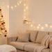 Petit salon lumineux décoré pour Noël avec un sapin mural pliable en pin clair, un canapé beige, une guirlande lumineuse chaude et des boules rouges mates.